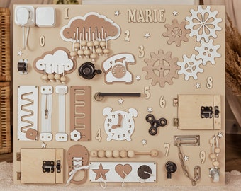 Peut inclure: Un tableau d'activités en bois beige avec différentes textures et activités pour les enfants. Il comprend une horloge, un téléphone, une fermeture éclair, un loquet, une chaîne, un labyrinthe, un curseur, un spinner, un ensemble d'engrenages et un ensemble de perles. Le tableau est étiqueté avec le nom "MARIE" et les chiffres de 0 à 9.