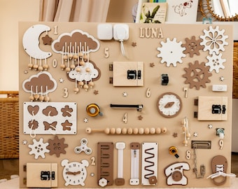 Könnte beinhalten: Ein beiges Busy Board aus Holz mit verschiedenen interaktiven Elementen, darunter weiße und braune Zahnräder, eine Mond- und Wolkenform mit hängenden Perlen, eine Uhr, ein Telefon, ein Schuh mit Schnürsenkeln, Holzzahlen und verschiedene Riegel.