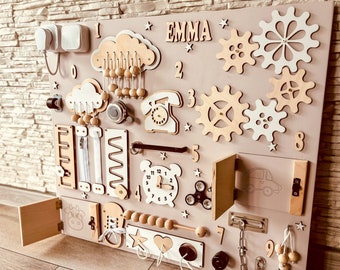 May include: A beige wooden activity board with various textures and activities for children. It features a cloud with beads, a phone, a clock, a door with a latch, a car, gears, a slider, a maze, and a chain. The board is labeled with numbers 1 through 9 and the name "EMMA".