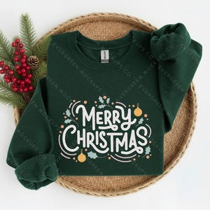 May include: A dark green long-sleeve sweatshirt featuring the white text Merry Christmas in a cursive script, accented with small light-coloured holly leaves and round ornaments. The garment is displayed on a round woven tray with pine branches.
