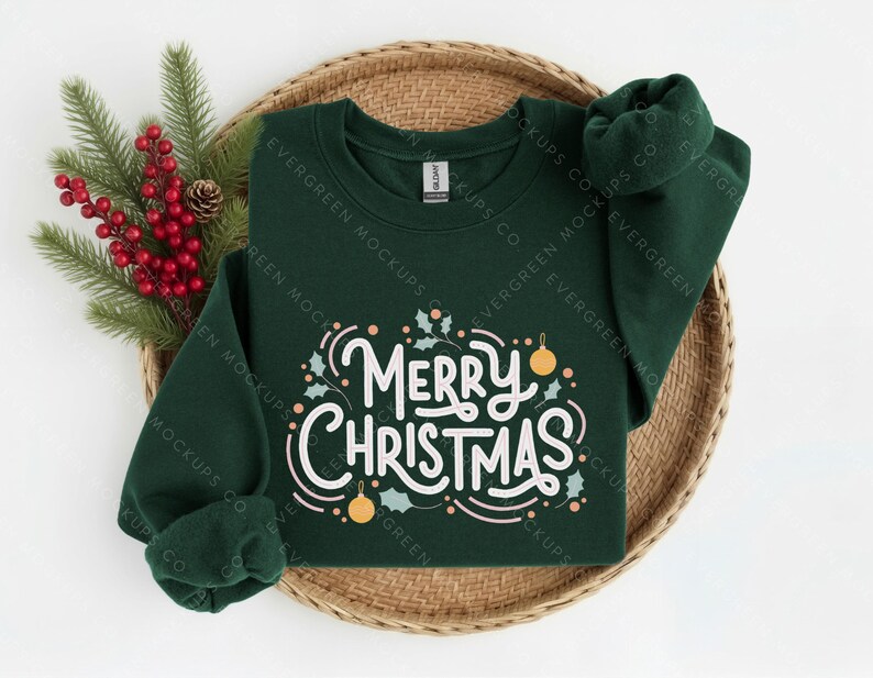 May include: A dark green long-sleeve sweatshirt featuring the white text Merry Christmas in a cursive script, accented with small light-coloured holly leaves and round ornaments. The garment is displayed on a round woven tray with pine branches.