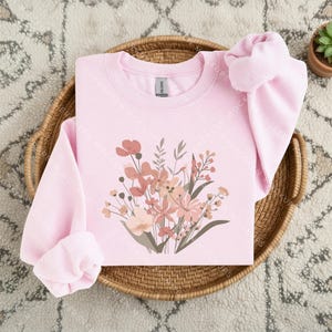 May include: A light pink long-sleeve crewneck sweatshirt featuring a central graphic print of a wildflower bouquet in shades of muted pink, peach, and brown. The sweatshirt has a ribbed collar, cuffs, and hem, with a visible brand label at the neck.
