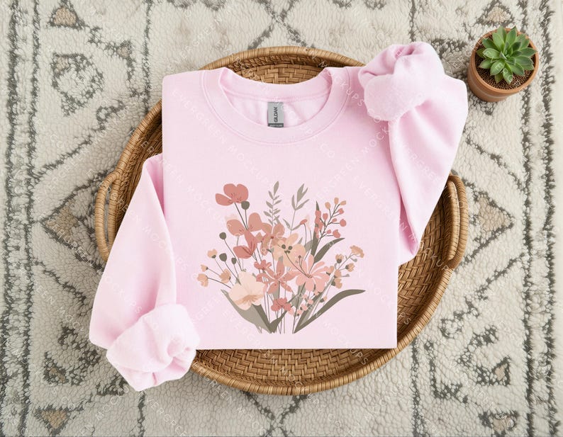 May include: A light pink long-sleeve crewneck sweatshirt featuring a central graphic print of a wildflower bouquet in shades of muted pink, peach, and brown. The sweatshirt has a ribbed collar, cuffs, and hem, with a visible brand label at the neck.