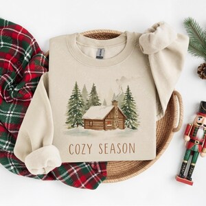 Pode incluir: Um moletom bege com uma ilustra&ccedil;&atilde;o em aquarela de uma cabana em uma floresta nevada, com o texto "COZY SEASON". Uma manta xadrez vermelha e verde, pinhas e um soldado de brinquedo tamb&eacute;m est&atilde;o na imagem.