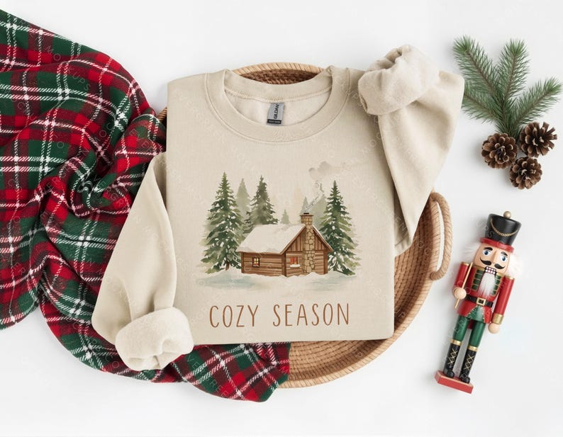 Pode incluir: Um moletom bege com uma ilustra&ccedil;&atilde;o em aquarela de uma cabana em uma floresta nevada, com o texto "COZY SEASON". Uma manta xadrez vermelha e verde, pinhas e um soldado de brinquedo tamb&eacute;m est&atilde;o na imagem.