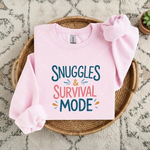 May include: A light pink long-sleeve sweatshirt featuring the dark blue and coral text "SNUGGLES & SURVIVAL MODE" printed on the front. The design includes small decorative dashes in blue and orange surrounding the ampersand and the word mode.