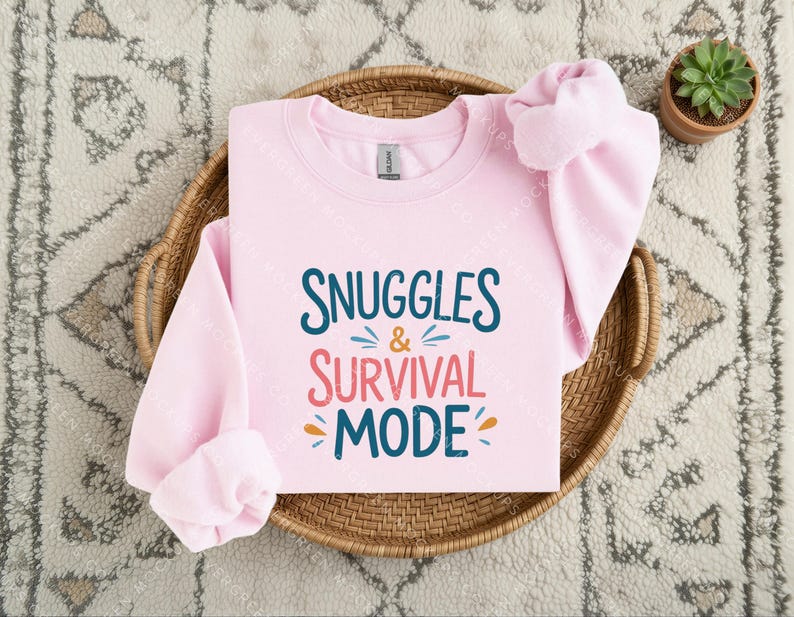 May include: A light pink long-sleeve sweatshirt featuring the dark blue and coral text "SNUGGLES & SURVIVAL MODE" printed on the front. The design includes small decorative dashes in blue and orange surrounding the ampersand and the word mode.
