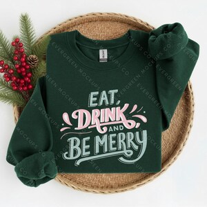 May include: A dark green long-sleeve sweatshirt featuring the text EAT, DRINK AND BE MERRY in a stylised font. The word DRINK is in light pink, while EAT, AND, and BE MERRY are in a light teal colour. The garment has a crew neckline.