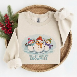 May include: A beige crewneck sweatshirt featuring a graphic of three snowmen wearing colorful hats and scarves. The snowmen are positioned above the blue text that reads "CHILLIN' WITH MY SNOWMIES." The garment has long sleeves and a ribbed neckline.