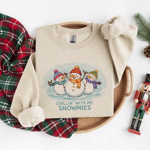 Pode incluir: Um moletom bege com um gr&aacute;fico de tr&ecirc;s bonecos de neve usando chap&eacute;us e cachec&oacute;is. O texto "Chillin' with my Snowmies" est&aacute; abaixo. O moletom est&aacute; em uma cesta tecida com um cobertor xadrez vermelho e verde.