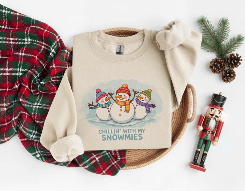 Pode incluir: Um moletom bege com um gr&aacute;fico de tr&ecirc;s bonecos de neve usando chap&eacute;us e cachec&oacute;is. O texto "Chillin' with my Snowmies" est&aacute; abaixo. O moletom est&aacute; em uma cesta tecida com um cobertor xadrez vermelho e verde.