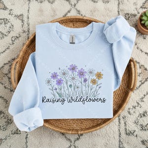 Peut inclure: Un sweat-shirt bleu clair &agrave; col rond orn&eacute; d'un motif de fleurs sauvages violettes, blanches et jaunes avec des tiges vertes. Le texte "Raising Wildflowers" est imprim&eacute; en police cursive noire sous l'illustration florale sur le devant.