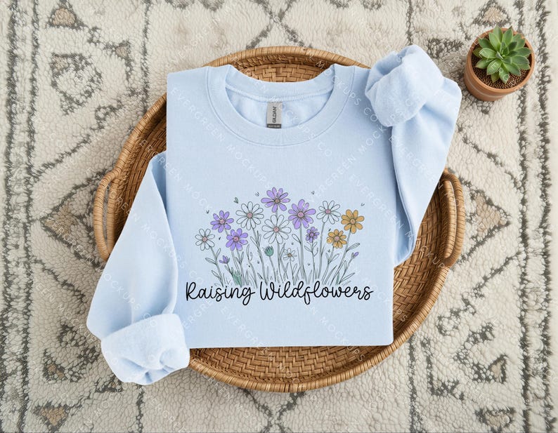 Peut inclure: Un sweat-shirt bleu clair &agrave; col rond orn&eacute; d'un motif de fleurs sauvages violettes, blanches et jaunes avec des tiges vertes. Le texte "Raising Wildflowers" est imprim&eacute; en police cursive noire sous l'illustration florale sur le devant.