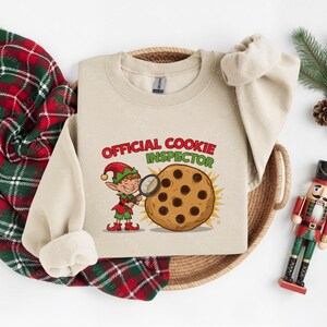 Pode incluir: Uma camisola bege com as palavras "OFFICIAL COOKIE INSPECTOR" em vermelho e verde. Um elfo com uma lupa examina um biscoito com pepitas de chocolate. Uma manta xadrez vermelha e verde e um quebra-nozes de brinquedo tamb&eacute;m est&atilde;o na imagem.