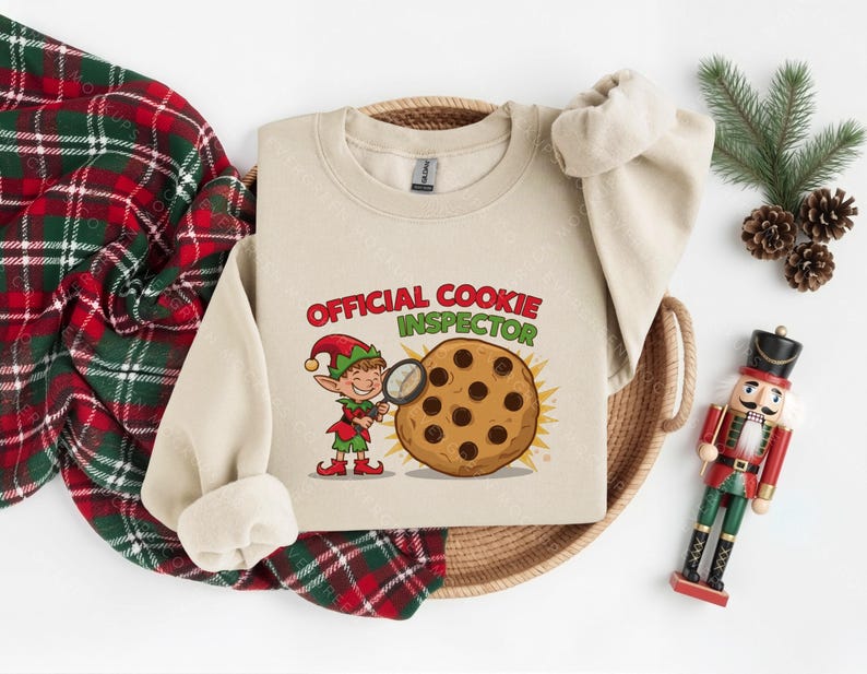 Pode incluir: Uma camisola bege com as palavras "OFFICIAL COOKIE INSPECTOR" em vermelho e verde. Um elfo com uma lupa examina um biscoito com pepitas de chocolate. Uma manta xadrez vermelha e verde e um quebra-nozes de brinquedo tamb&eacute;m est&atilde;o na imagem.