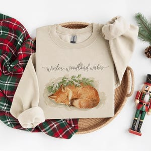 Pode incluir: Uma camisola bege com o texto "Winter woodland wishes" e uma ilustra&ccedil;&atilde;o em aquarela de uma raposa dormindo. A camisola est&aacute; em uma cesta tecida, com um cobertor xadrez vermelho e verde, pinhas e um soldado de brinquedo.