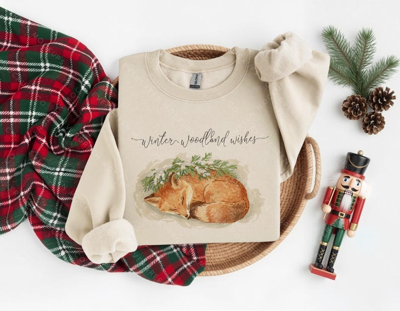 Pode incluir: Uma camisola bege com o texto "Winter woodland wishes" e uma ilustra&ccedil;&atilde;o em aquarela de uma raposa dormindo. A camisola est&aacute; em uma cesta tecida, com um cobertor xadrez vermelho e verde, pinhas e um soldado de brinquedo.