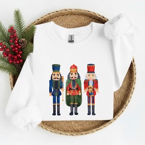 May include: White sweatshirt featuring three watercolor nutcracker figures in blue, green, and red uniforms. The nutcrackers have black boots and hats. The sweatshirt is displayed on a woven basket with red berries and pine needles.