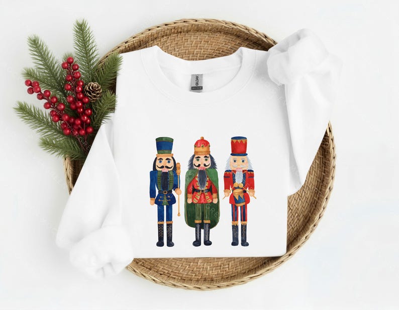 May include: White sweatshirt featuring three watercolor nutcracker figures in blue, green, and red uniforms. The nutcrackers have black boots and hats. The sweatshirt is displayed on a woven basket with red berries and pine needles.