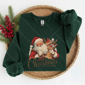 May include: A dark green long-sleeved sweatshirt featuring a graphic of Santa Claus holding a sack filled with gifts. Below the illustration, the text reads A VERY MERRY Christmas in a stylised script font. The garment has a crew neckline.