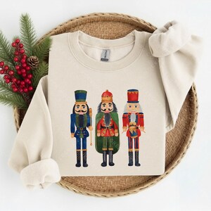 May include: A sand-colored crewneck sweatshirt featuring a graphic print of three nutcracker figures. The left nutcracker wears a blue uniform, the center one wears a red and green uniform with a crown, and the right one wears a red uniform.