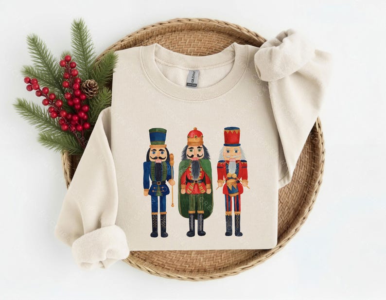 May include: A sand-colored crewneck sweatshirt featuring a graphic print of three nutcracker figures. The left nutcracker wears a blue uniform, the center one wears a red and green uniform with a crown, and the right one wears a red uniform.