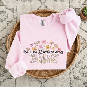 May include: A light pink crewneck sweatshirt featuring a graphic of colourful wildflowers in shades of yellow, pink, and orange. The text "Raising Wildflowers" is printed in a black cursive font across the centre of the floral illustration.