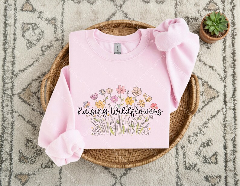 May include: A light pink crewneck sweatshirt featuring a graphic of colourful wildflowers in shades of yellow, pink, and orange. The text "Raising Wildflowers" is printed in a black cursive font across the centre of the floral illustration.