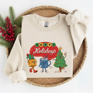 May include: A beige crewneck sweatshirt featuring a graphic print with the text Happy Holidays in a red speech bubble. The design includes a cartoon gift box, a blue mug with a face, and a decorated Christmas tree with arms and legs.