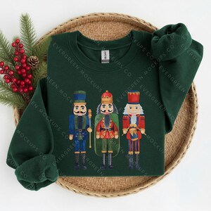 May include: A dark green long-sleeve sweatshirt featuring a graphic print of three nutcracker figures in blue, red, and green uniforms. The centre nutcracker wears a crown, while the others wear tall hats. The sweatshirt has a crew neckline.