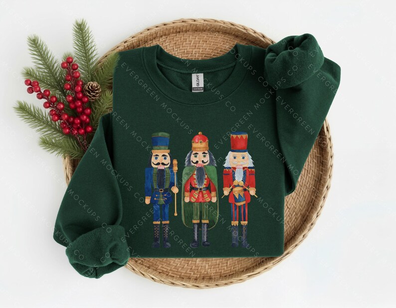 May include: A dark green long-sleeve sweatshirt featuring a graphic print of three nutcracker figures in blue, red, and green uniforms. The centre nutcracker wears a crown, while the others wear tall hats. The sweatshirt has a crew neckline.