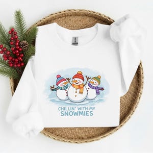 May include: White sweatshirt featuring a cartoon illustration of three snowmen with colorful hats and scarves. The text "Chillin' with my Snowmies" is below the snowmen. The sweatshirt is displayed on a woven tray with decorative greenery.