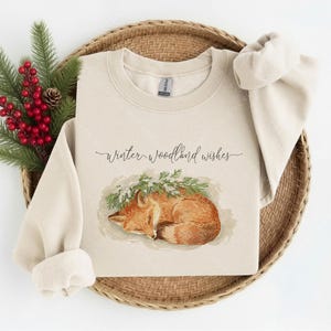 May include: A sand-colored crewneck sweatshirt featuring a graphic of a sleeping orange fox nestled under green pine branches. The text "winter woodland wishes" is printed in a dark cursive script above the fox illustration on the front.