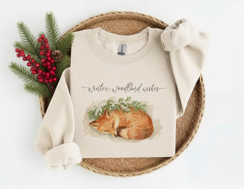 May include: A sand-colored crewneck sweatshirt featuring a graphic of a sleeping orange fox nestled under green pine branches. The text "winter woodland wishes" is printed in a dark cursive script above the fox illustration on the front.