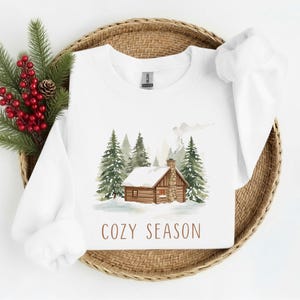 May include: White sweatshirt with a watercolor illustration of a cabin in a snowy forest. The phrase "COZY SEASON" is printed in brown below the image. The sweatshirt is displayed on a woven basket with evergreen branches and red berries.