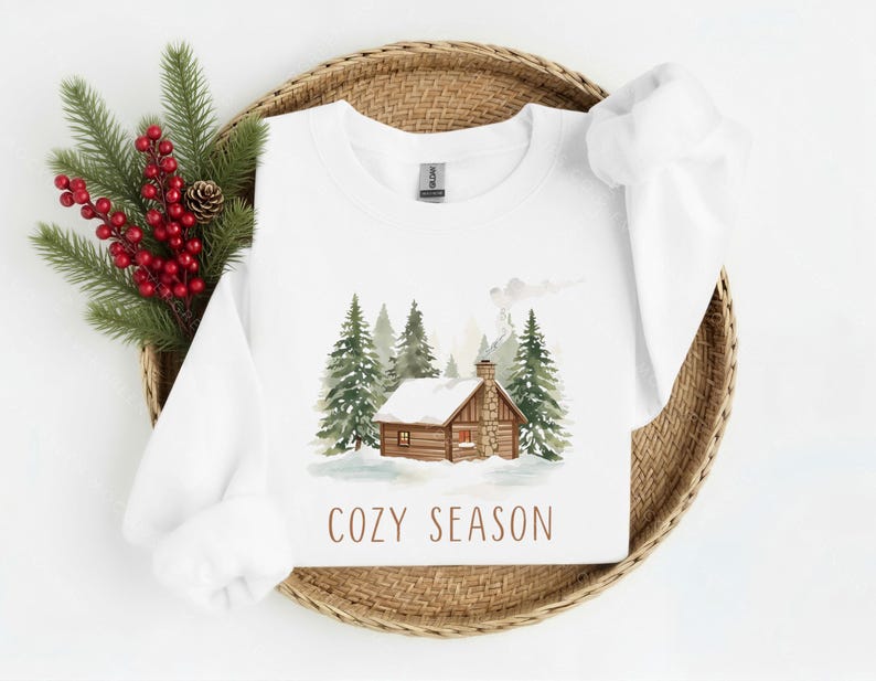 May include: White sweatshirt with a watercolor illustration of a cabin in a snowy forest. The phrase "COZY SEASON" is printed in brown below the image. The sweatshirt is displayed on a woven basket with evergreen branches and red berries.