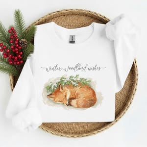 May include: White sweatshirt with a watercolor illustration of a sleeping fox, surrounded by greenery. The text "Winter woodland wishes" is written in cursive above the fox. The sweatshirt is displayed on a woven basket with red berries and pine branches.