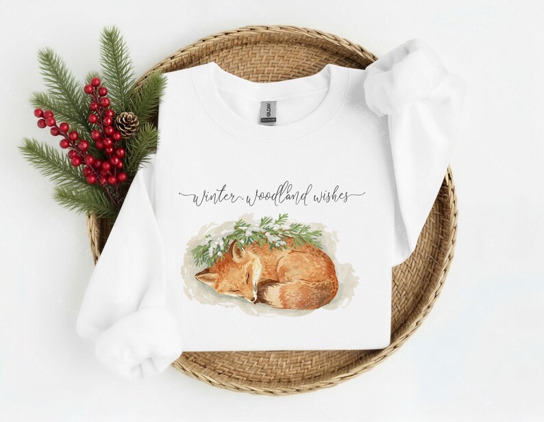 May include: White sweatshirt with a watercolor illustration of a sleeping fox, surrounded by greenery. The text "Winter woodland wishes" is written in cursive above the fox. The sweatshirt is displayed on a woven basket with red berries and pine branches.