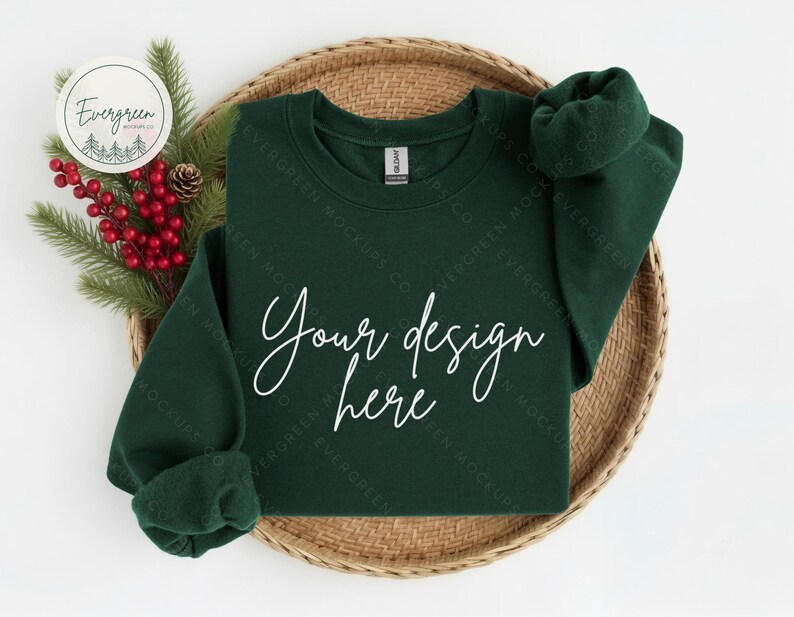 May include: A dark forest green crewneck sweatshirt featuring the white text Your design here printed on the front. The garment is displayed on a woven circular tray alongside a decorative sprig of pine needles and red berries.