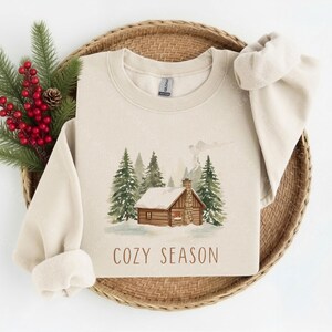 May include: A sand-colored crewneck sweatshirt featuring a watercolor-style graphic of a log cabin nestled among snow-covered pine trees. The text "COZY SEASON" is printed in brown lettering below the cabin illustration on the front of the garment.