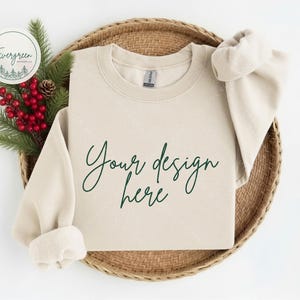 May include: A sand-colored crewneck sweatshirt featuring the dark green cursive text Your design here printed on the front. The garment has long sleeves with ribbed cuffs and a ribbed neckline, displayed with a sprig of pine and red berries.