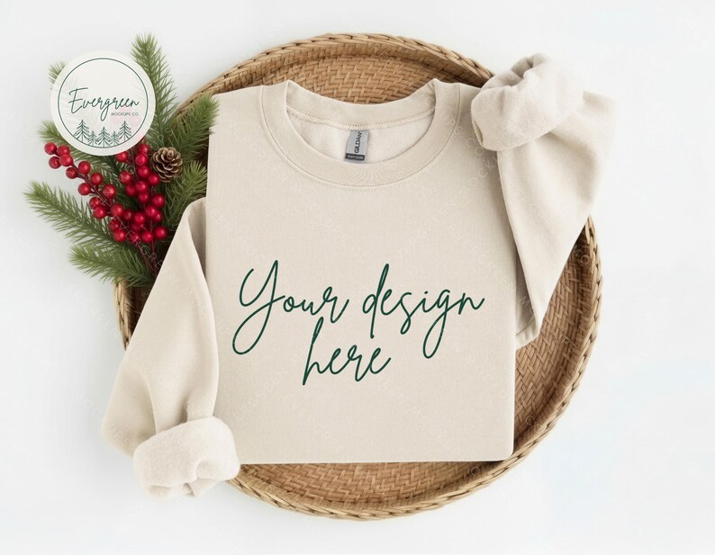 May include: A sand-colored crewneck sweatshirt featuring the dark green cursive text Your design here printed on the front. The garment has long sleeves with ribbed cuffs and a ribbed neckline, displayed with a sprig of pine and red berries.