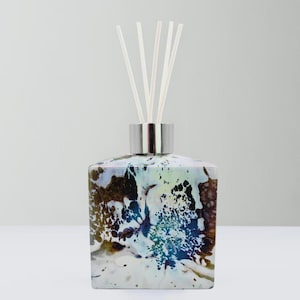 May include: A decorative reed diffuser with a square, iridescent glass bottle. The bottle has a marbled pattern in brown, blue, and white. It features a silver-coloured metal collar and several white reeds.