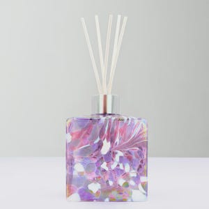 May include: A decorative glass diffuser bottle with a silver collar and white reeds. The bottle has a square shape and is decorated with a colourful, abstract pattern in shades of purple, pink, and white.