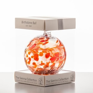 May include: A clear glass bauble with a red and white floral design. The bauble is packaged in a clear box with a white label that reads "Birthstone Ball JULY - RUBY" and "The Sienna Collection".