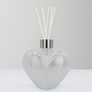 May include: An iridescent heart-shaped glass diffuser bottle with a silver collar and white reeds. The bottle has a pearlescent finish, reflecting a rainbow of colours. A decorative home fragrance item.
