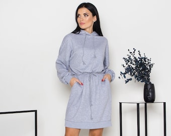May include: A gray hooded sweatshirt dress with a drawstring waist. The dress has long sleeves and two pockets. The model is wearing the dress and standing in front of a white wall.