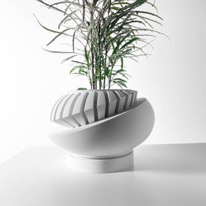 May include: A contemporary white plant pot with a distinctive geometric design. It has a spherical base and a top section with vertical, angled grooves. Green leafy plant stems emerge from the top. The pot is a stylish home decor item.
