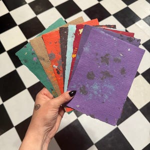 May include: A fan of rectangular handmade paper sheets with deckled edges in various colors including purple, red, orange, teal, and tan, featuring speckled textures and metallic flecks throughout the surface of each individual paper sheet.