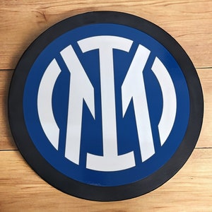 Könnte beinhalten: Eine runde Wanddekoration mit schwarzem Rahmen und einem blauen Kreis in der Mitte. Der Kreis zeigt ein weißes, stilisiertes "M"-Logo.